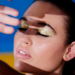 Close-up of a woman with her eyes closed, wearing metallic gold eyeshadow and a nude pink lip, with a hand shielding her forehead against a blue and yellow background.